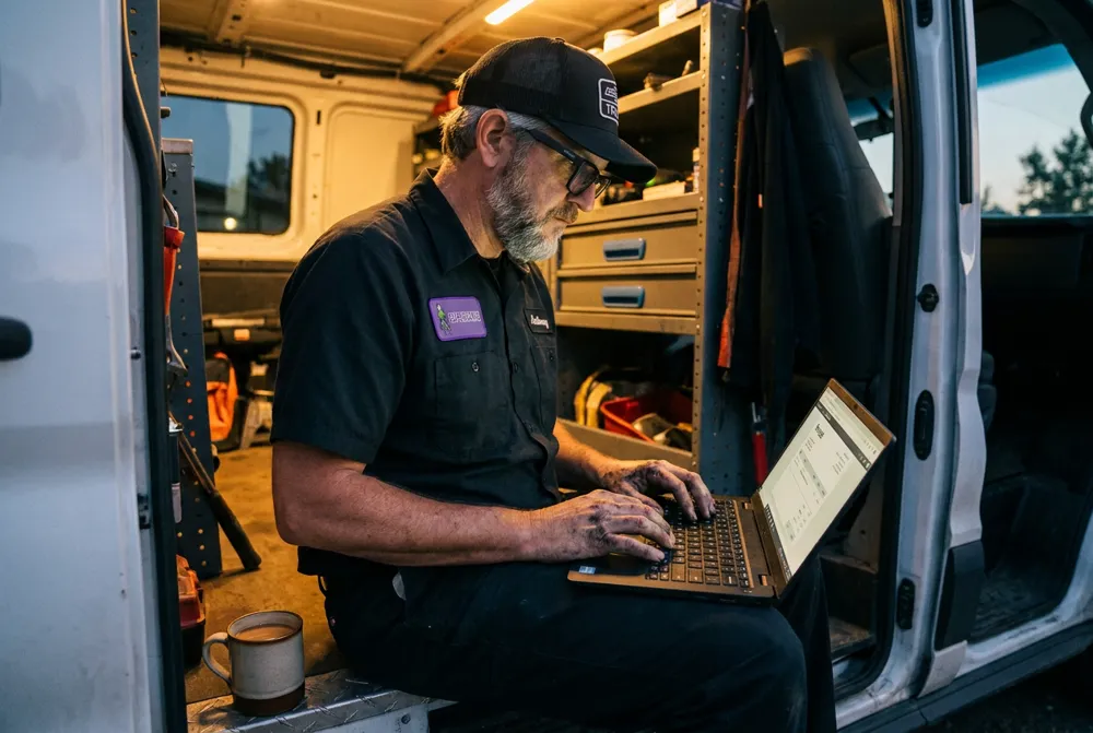 Anthony sitting in the side door of his van at dusk, sending an invoice from a laptop
